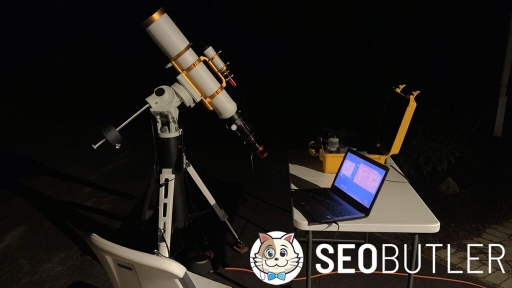 A nighttime outdoor setup for astrophotography featuring a white and gold refractor telescope on a tripod mount, aimed at the sky. In front of it is a white folding chair and a table holding an open laptop displaying astrophotography software, along with a yellow hard case and accessories. The scene is dimly lit, with shadows cast on the pavement.