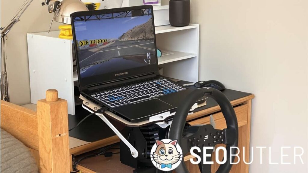 A sim racing setup on a wooden desk features a Predator gaming laptop displaying a desert racing game. A FANATEC racing wheel is securely mounted below the laptop, and a computer mouse sits beside it. The background includes a white shelf, a metallic desk lamp, and a wooden bedpost.
