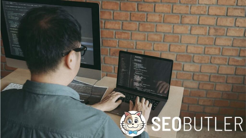 A software developer wearing glasses and a teal shirt is seated at a desk, working on a laptop and an external monitor displaying lines of code. The setup is against a brick wall background.