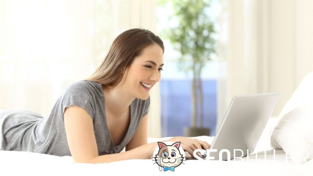 A young woman with light skin and long brown hair lies on a white bed, smiling while typing on a silver laptop. She wears a casual gray T-shirt, with soft natural light and blurred greenery in the background.