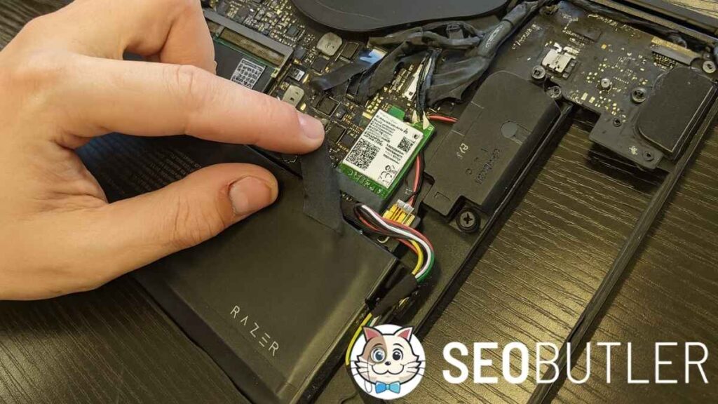 A person's hand lifts black tape inside a Razer laptop, revealing the installed WiFi card and internal wiring on a dark wooden surface.