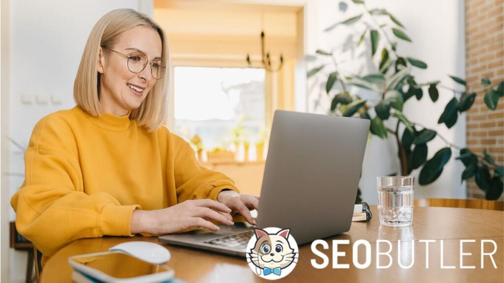 A smiling woman with blonde hair and glasses wearing a yellow sweater works on a laptop at a wooden table, with a notebook, mouse, glass of water, and indoor plants in the background.