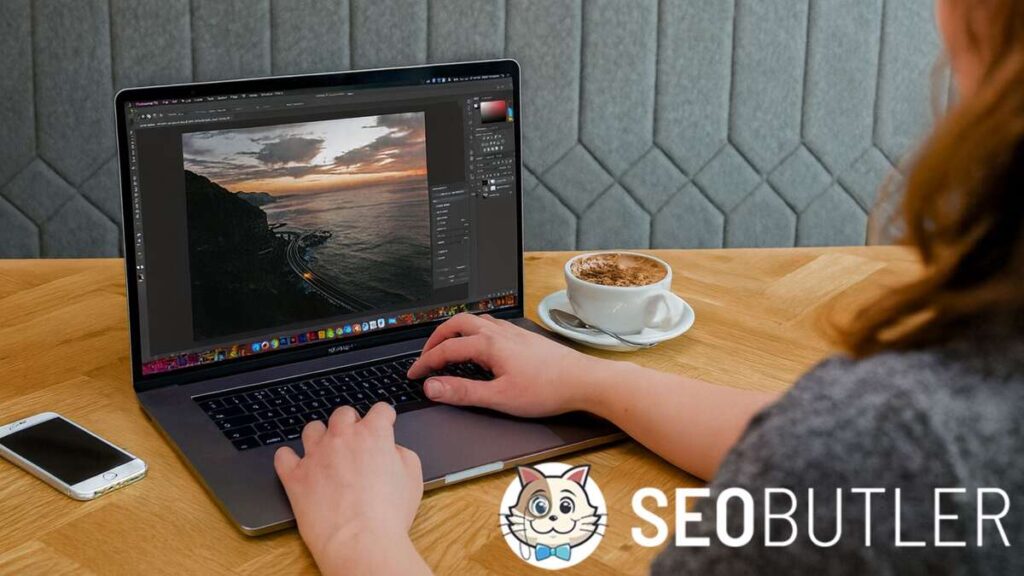 A woman is working on a MacBook Pro at a café table, editing a sunset coastal photo. A cappuccino and smartphone are placed nearby.