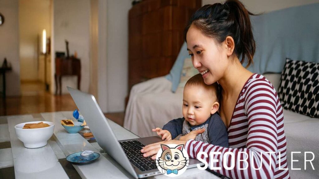 A mother sits on a sofa, smiling while working on a laptop with her baby on her lap. The baby looks at the screen in a cozy living room setting.
