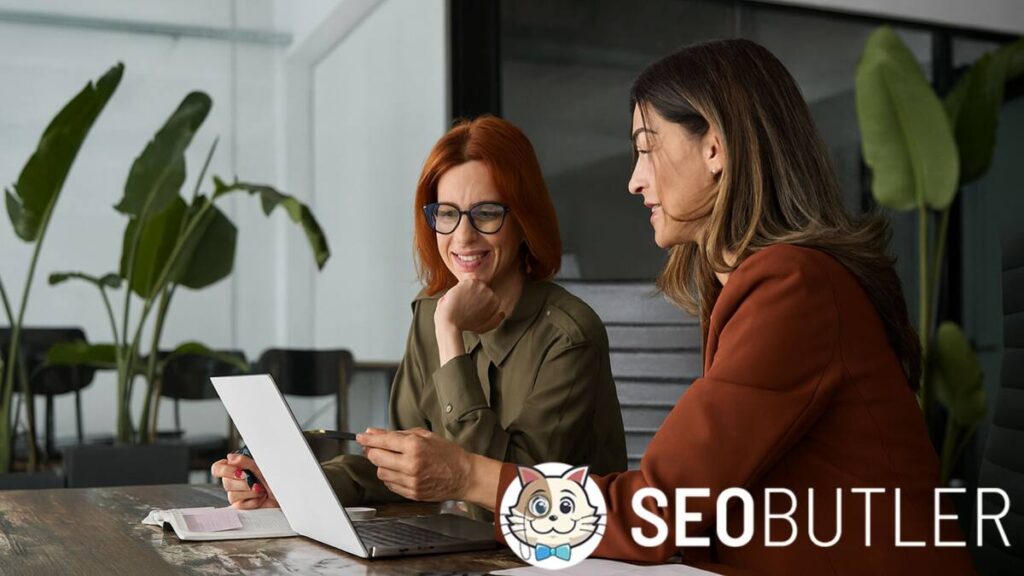 Two professional women sit at a desk in a modern office, smiling and discussing something on a laptop screen, with notebooks and plants in the background.