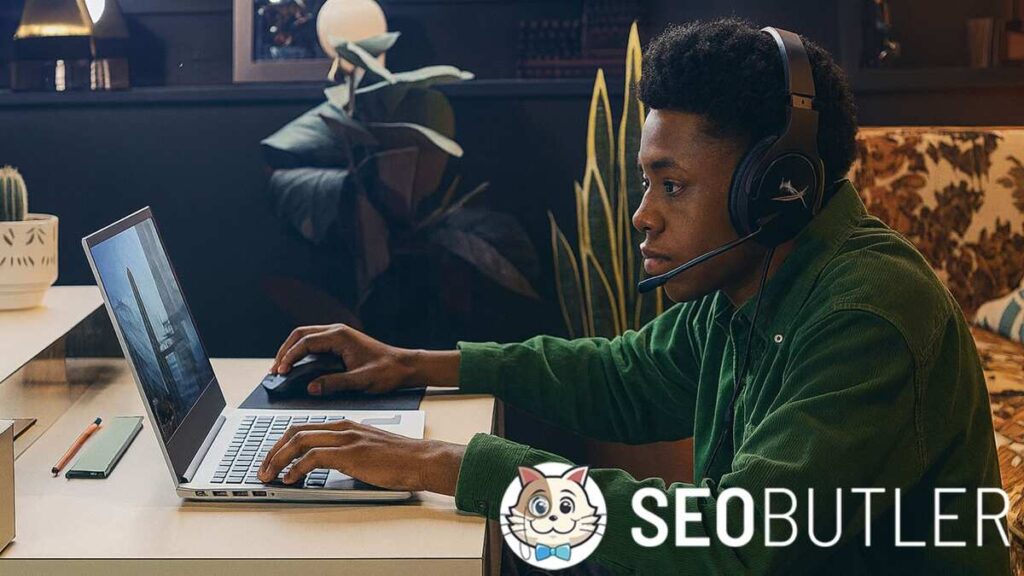 A young man wearing headphones with a mic, sitting at a desk and gaming or streaming on a silver laptop. He is dressed in a green shirt with short curly hair, focused on the screen in a cozy room with plants and warm decor.