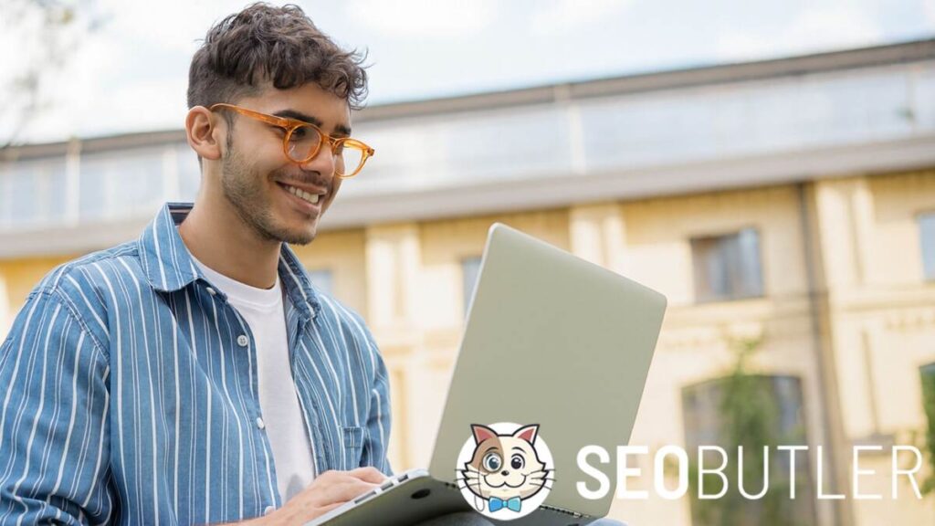 A young man wearing orange glasses and a blue striped shirt is smiling while using a laptop outdoors, with a modern building and trees in the background.