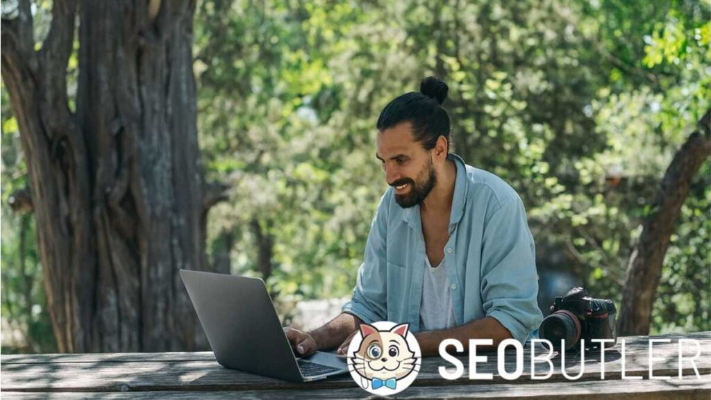 A man with a beard and tied-back hair sits outdoors at a wooden table, smiling while working on a laptop, with a camera placed beside him and trees in the background.