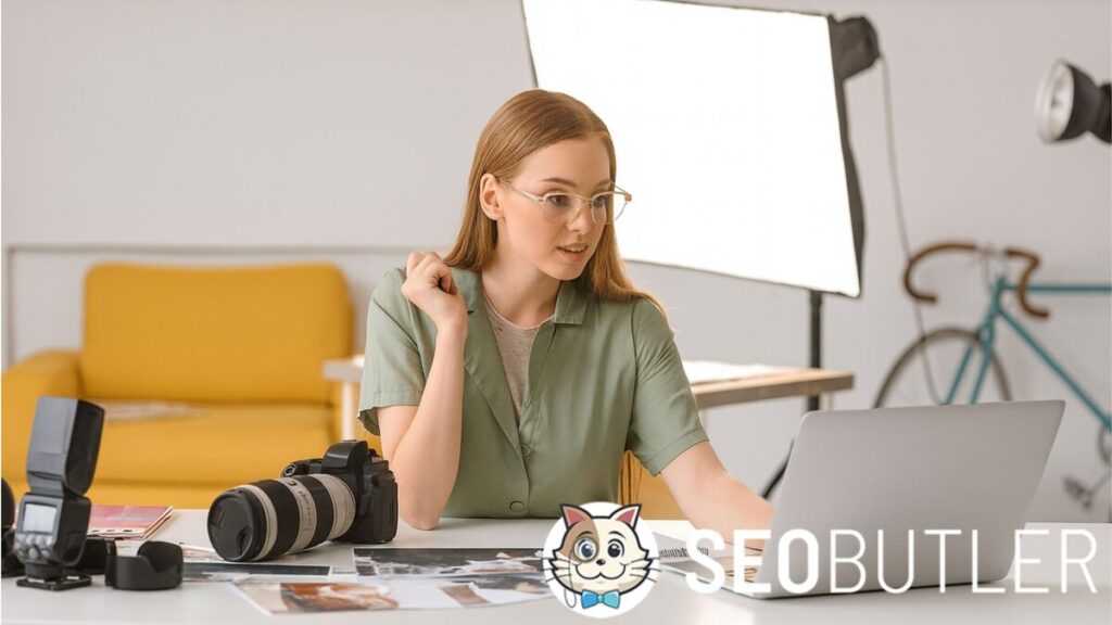 A young woman with long light brown hair and glasses sits at a desk in a bright studio, working on a laptop with a camera, photos, and lighting equipment around her.