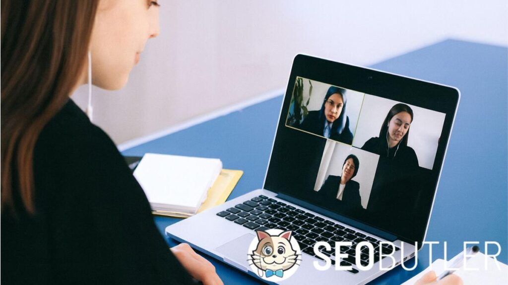 A woman sits at a desk with a laptop, participating in a video conference call with three other women displayed on the screen, while taking notes with a pen.