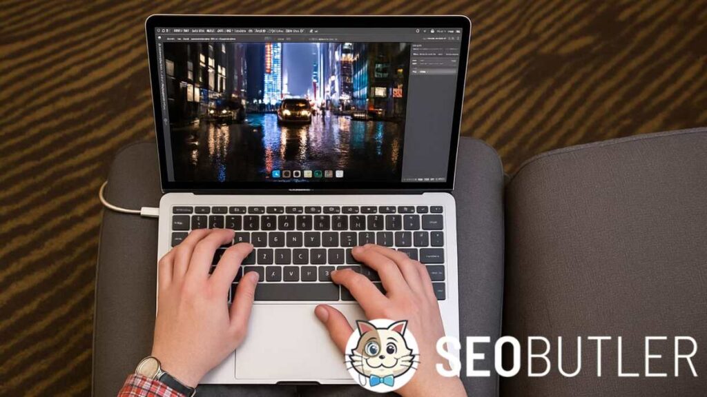 A person sitting on a gray armchair using a MacBook Pro on their lap. The laptop screen shows a city night scene being edited, while the person wears a red plaid shirt and types on the illuminated keyboard in a warmly lit room.