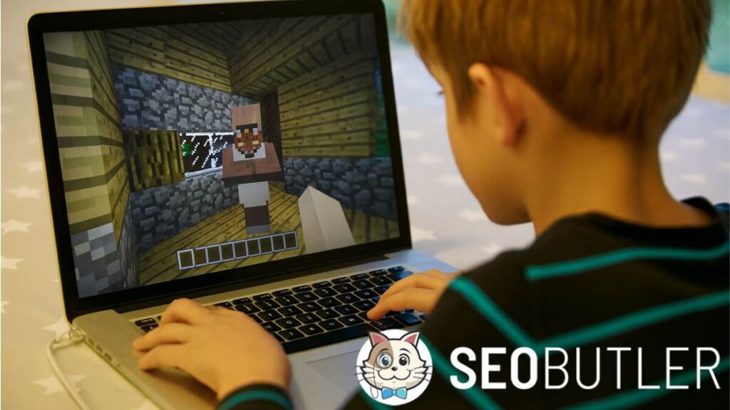 A young boy playing Minecraft on a laptop at a table with a star-patterned tablecloth, showing a villager inside a wooden house on the game screen.